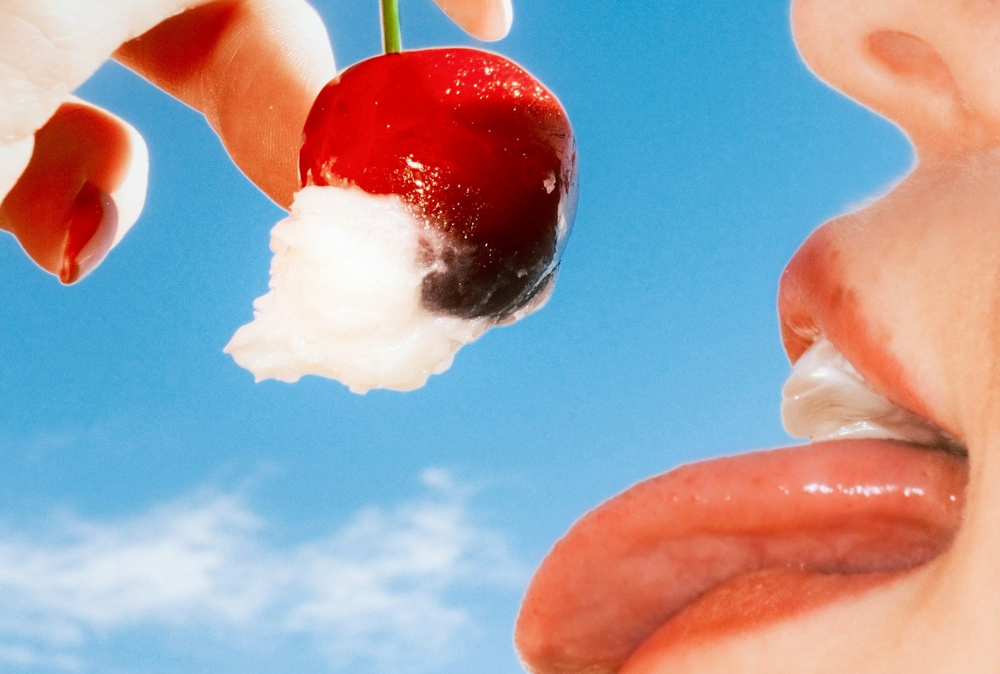 Hand offering cherry to mouth against blue sky